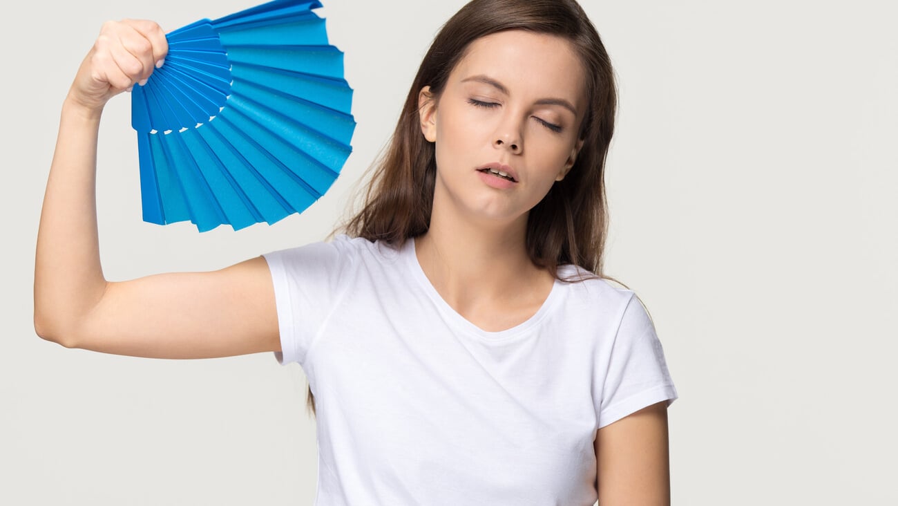 Tired young woman feel overheated suffering from heat stroke high temperature sweating problem, sweaty girl holding waving fan cooling in hot summer weather isolated on white grey studio background; Shutterstock ID 1325627747