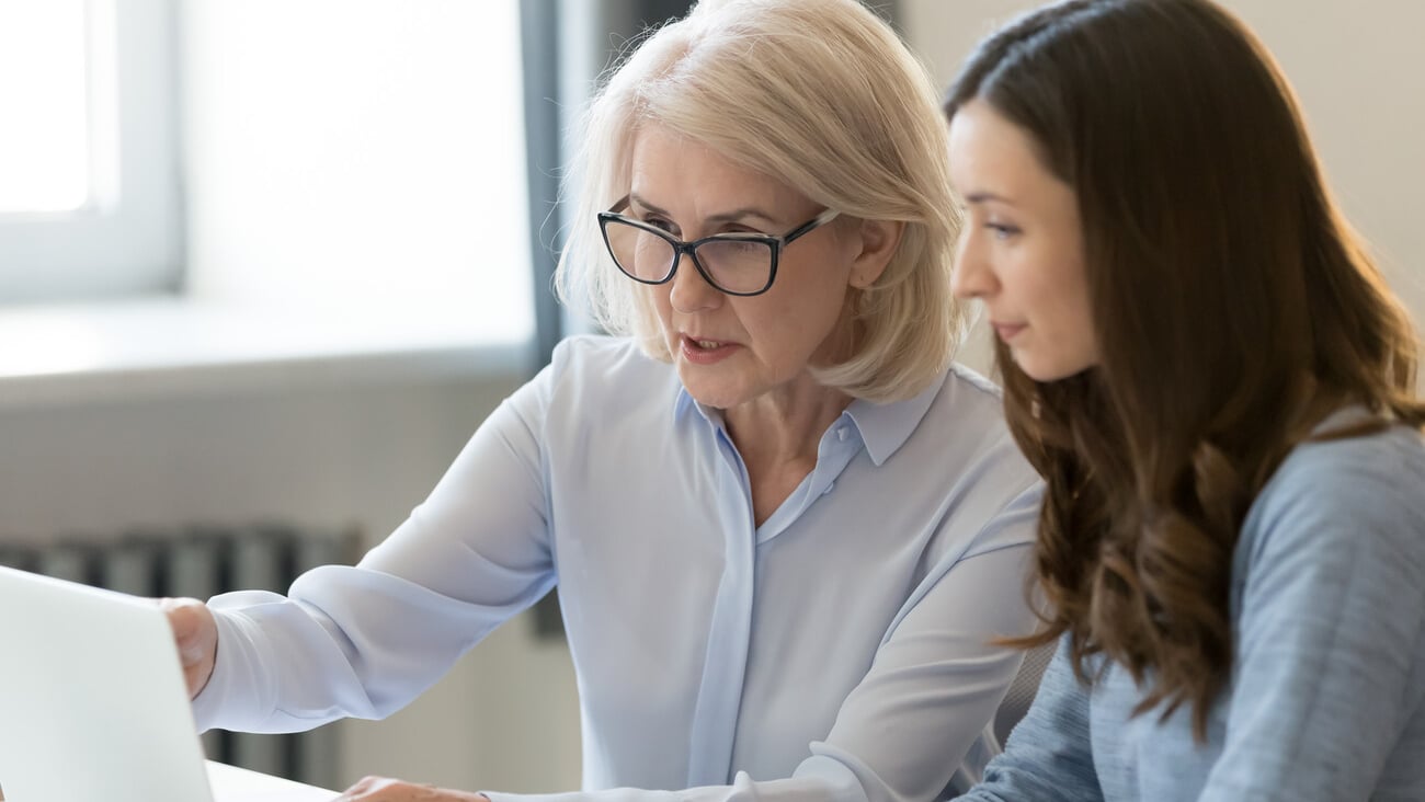 Serious old female mentor teacher coach teaching intern or student computer work pointing at laptop, mature executive manager explaining online project to young employee learning new skills in office; Shutterstock ID 1332825533