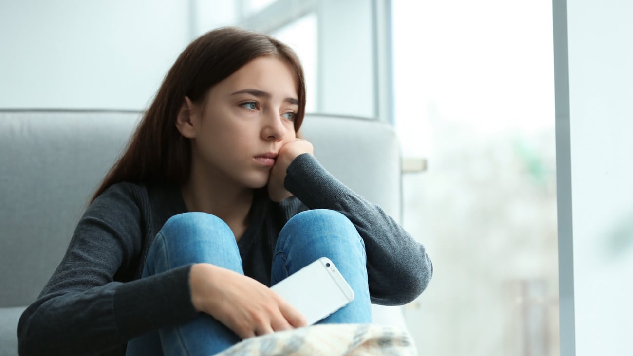 Upset teenage girl with smartphone sitting at window indoors. Space for text; Shutterstock ID 1373030828