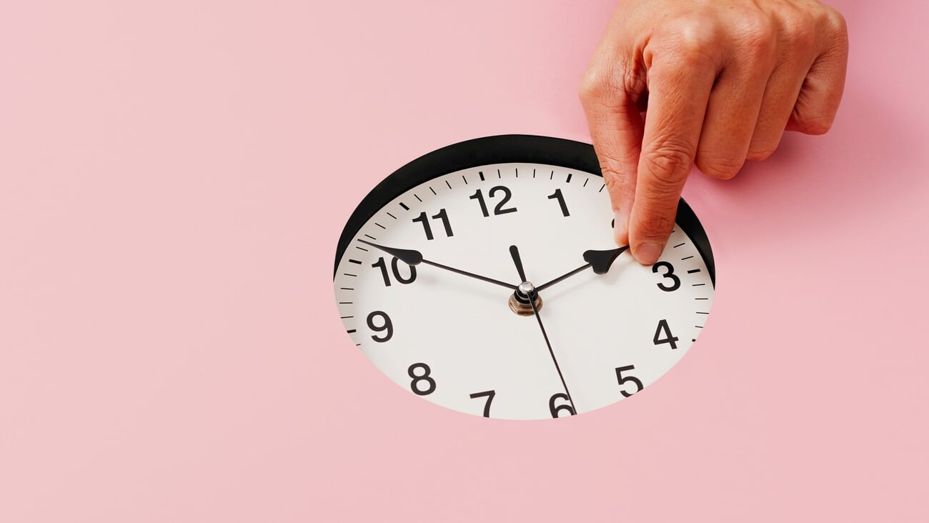 the hand of a young caucasian man sets a clock backward or forward, at the end or at the beginning of the summer, on a pink background; Shutterstock ID 2042342204
