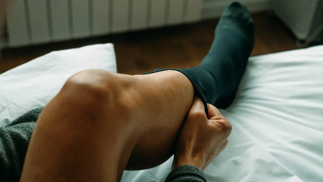 a man is putting on a compression sock sitting on his bed at home; Shutterstock ID 2260283603