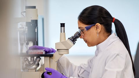 Une scientifique vêtue d'une blouse blanche et de lunettes de protection examine attentivement un échantillon à l'aide d'un microscope de laboratoire professionnel.