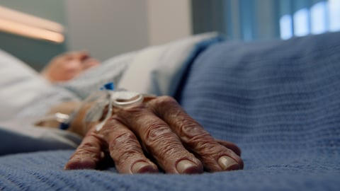 Gros plan sur la main d'un aîné, qui est couché dans un lit d'hôpital.