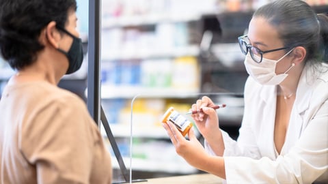Une pharmacienne conseille sa patiente au comptoir derrière une vitre en plexiglas 