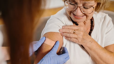 Un professionnel de santé applique un pansement sur le haut du bras d'une femme âgée souriante, portant des lunettes, après une vaccination.