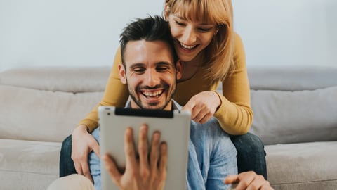 Happy couple of millennials using digital tablet at home. Boyfriend and girlfriend sitting on the sofa in the living room searching something on internet. People and technology concept.; Shutterstock ID 1819689368