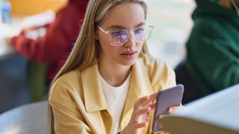 Teen girl gen z student using mobile phone looking at smartphone sitting at desk in university college campus classroom. Young blonde woman holding cellphone modern tech in university.; Shutterstock ID 2343942511