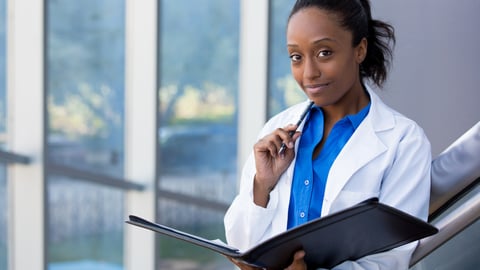 Closeup headshot portrait of friendly, smiling confident female doctor, healthcare professional with labcoat, holding pen to face and holding notebook pad. Isolated hospital clinic background.; Shutterstock ID 242631055